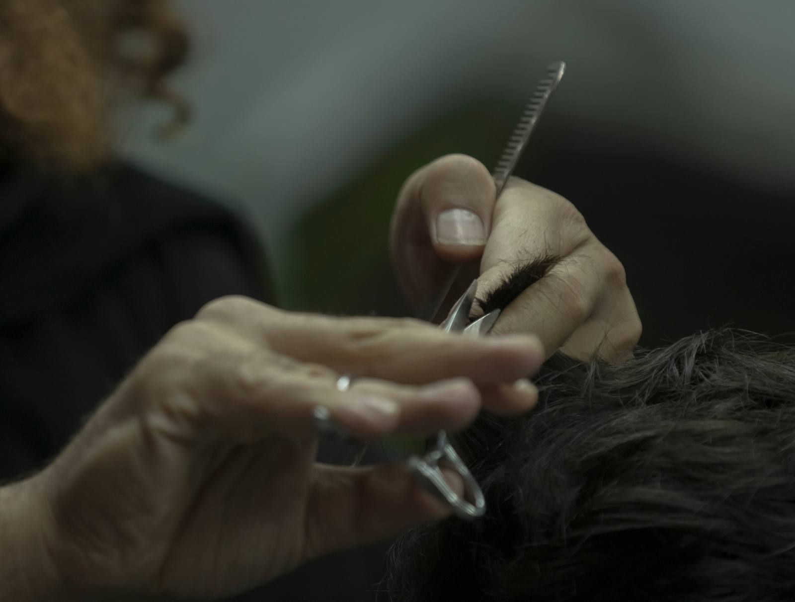 A barber's hands holding a section of dark hair between comb and scissors