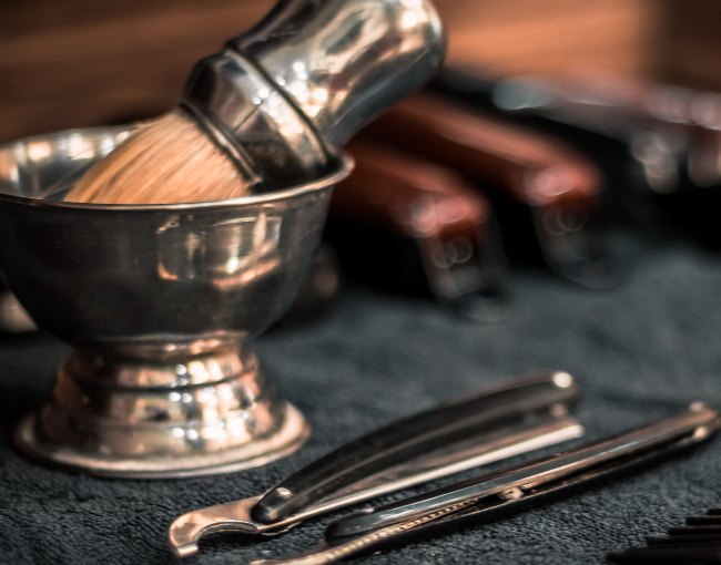 A shaving brush in a steel bowl beside two straight razors on a dark towel