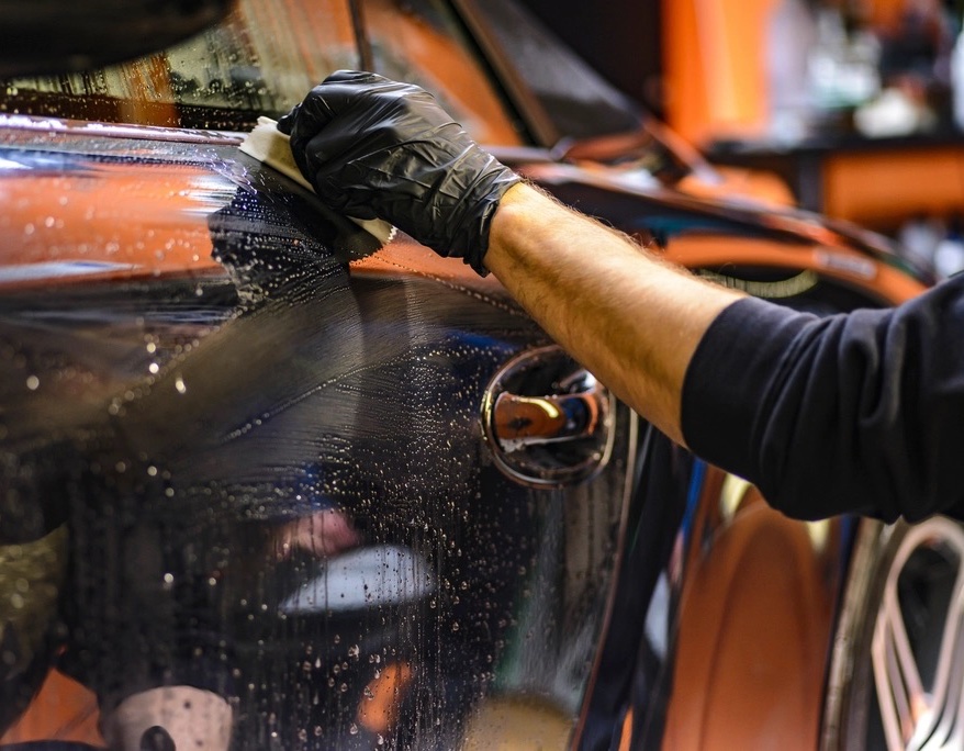 A gloved hand wiping soapy water across the dark, wet door of a car during a hand wash