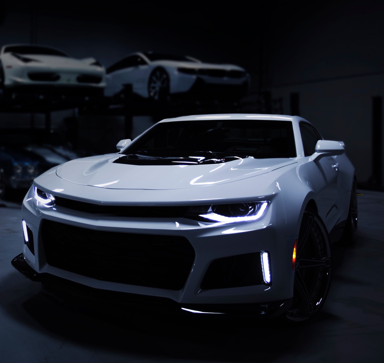 A white sports coupe with dark-tinted glass parked in a dim studio bay, more cars stacked on lifts behind it